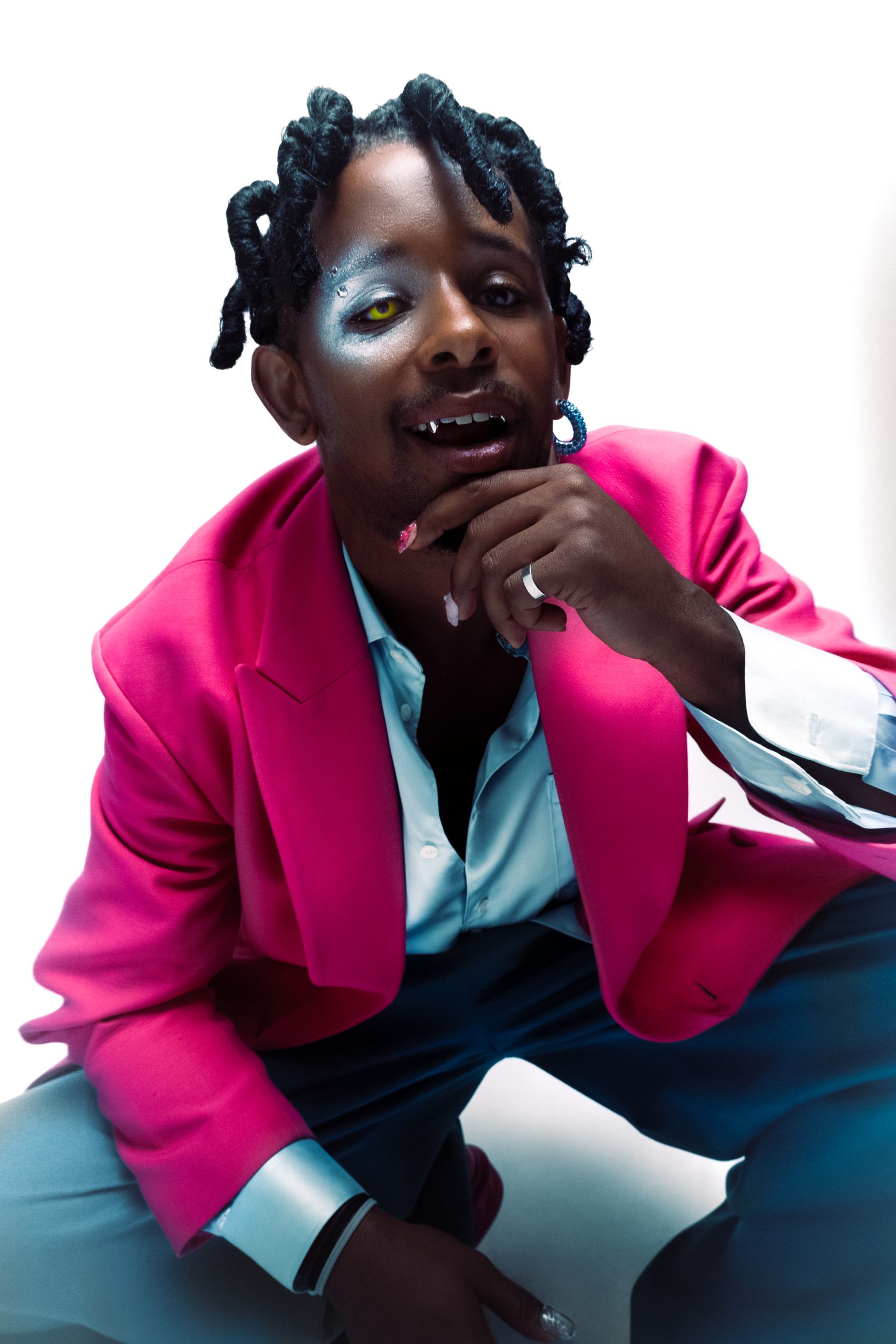 black model bending down in pink and blue suit with extravagant nails and shiny makeup