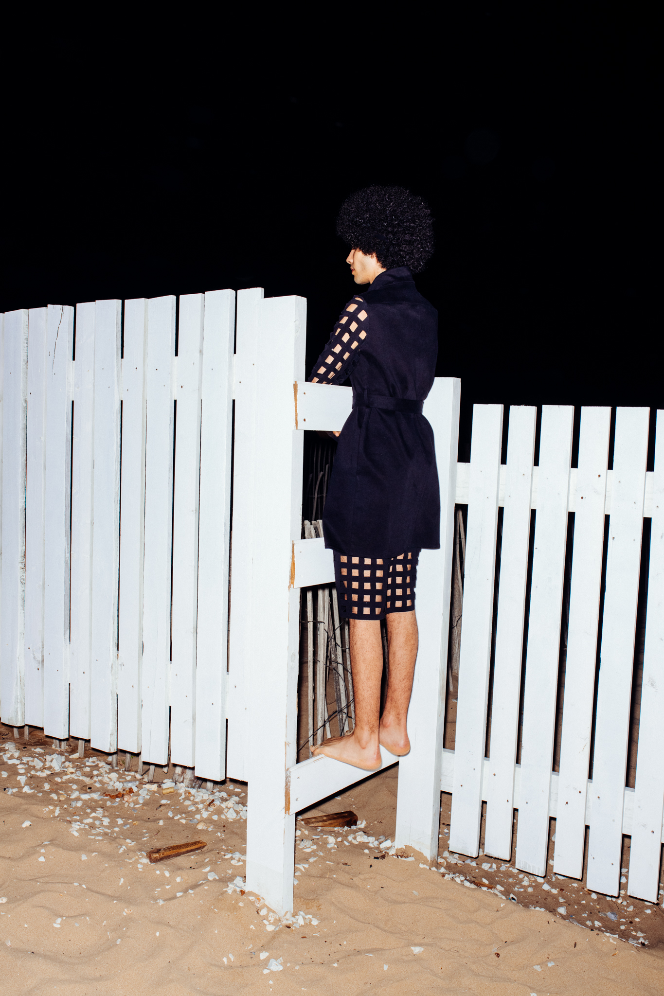 tunisian model Yassin Walhezi standing on a fence at the beach. He's wearing an outfit by Anissa Aida. Afro