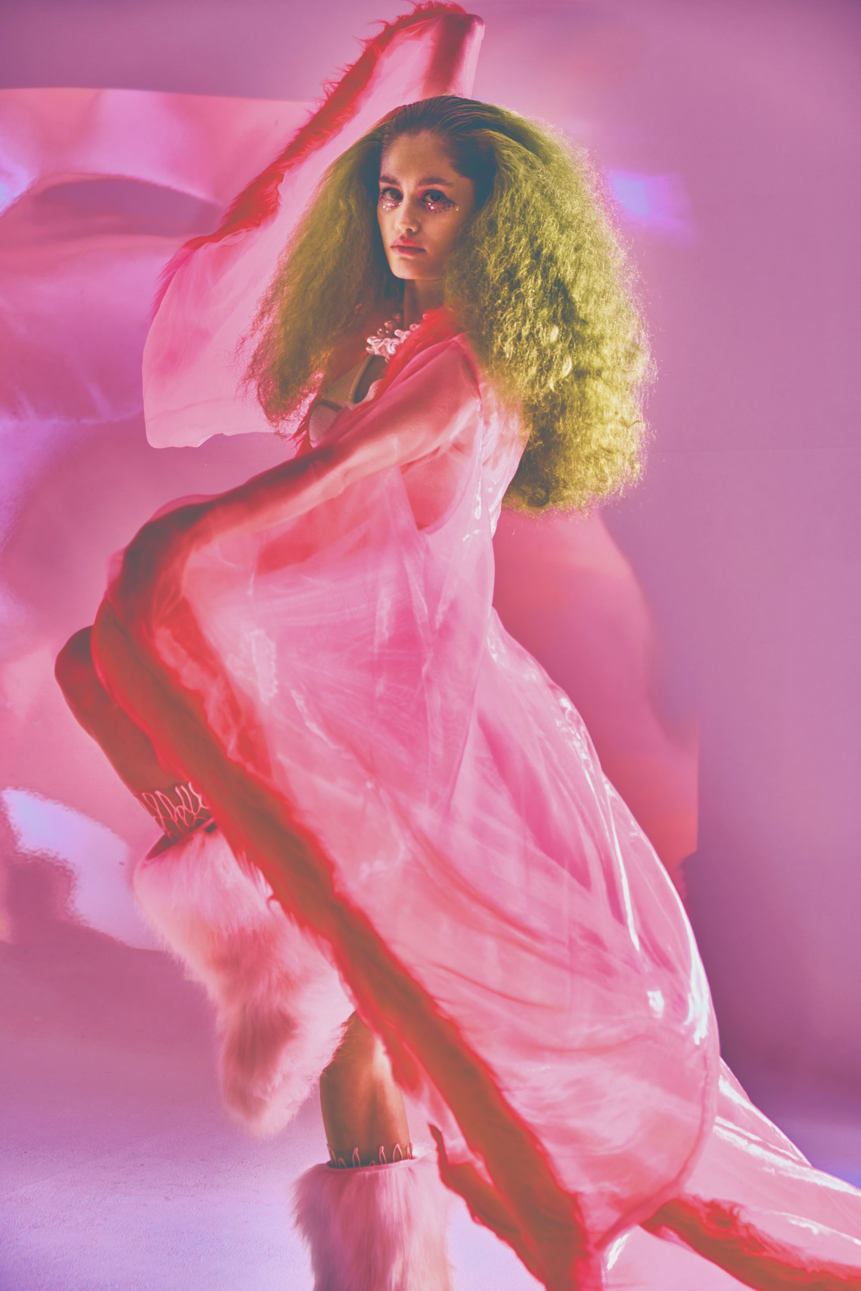 full body shot from the side of White Female Model in sheer pink cape and voluminous hair in front of pink blue background