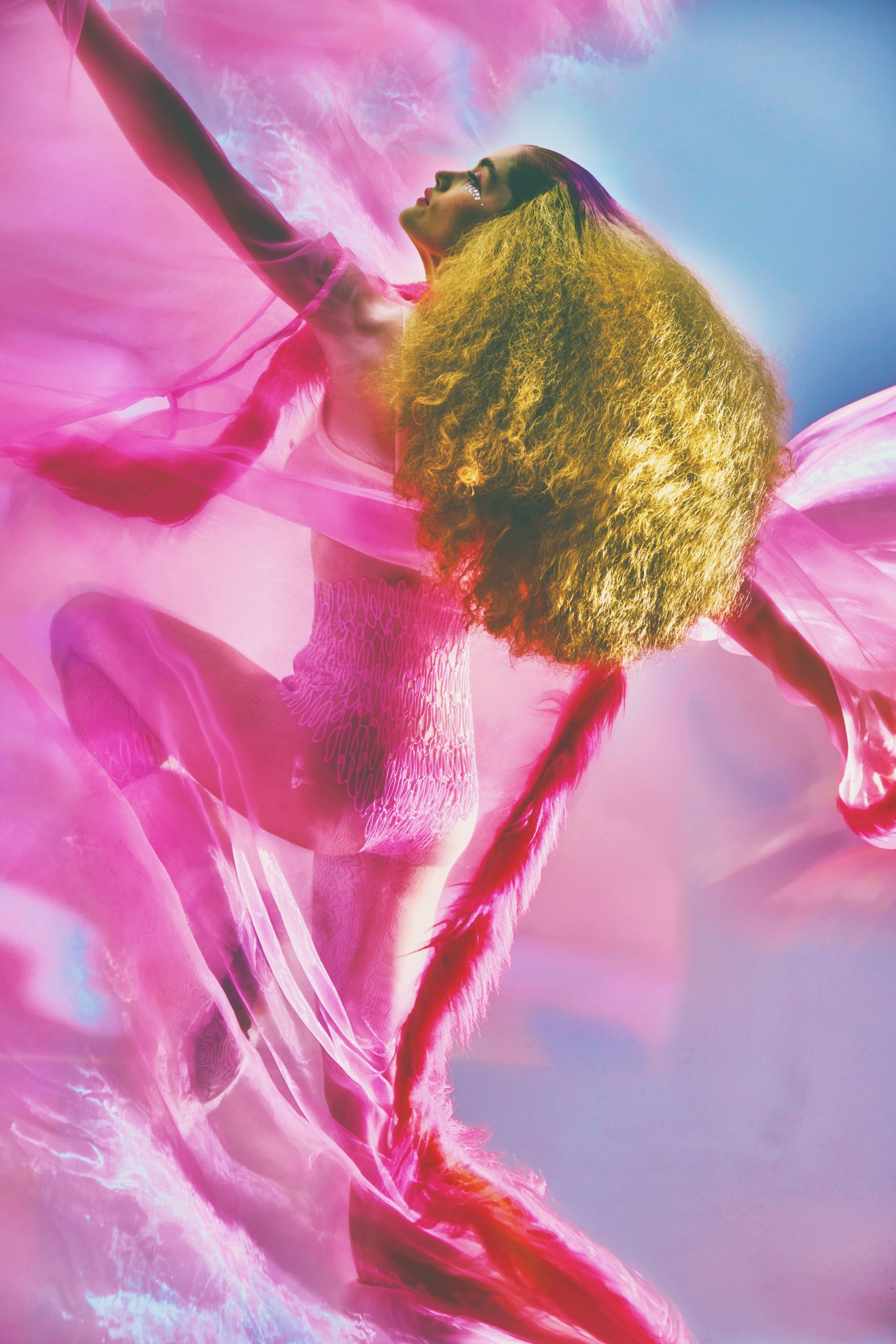 full body shot from the back of White Female Model in sheer pink cape and voluminous hair in front of pink blue background