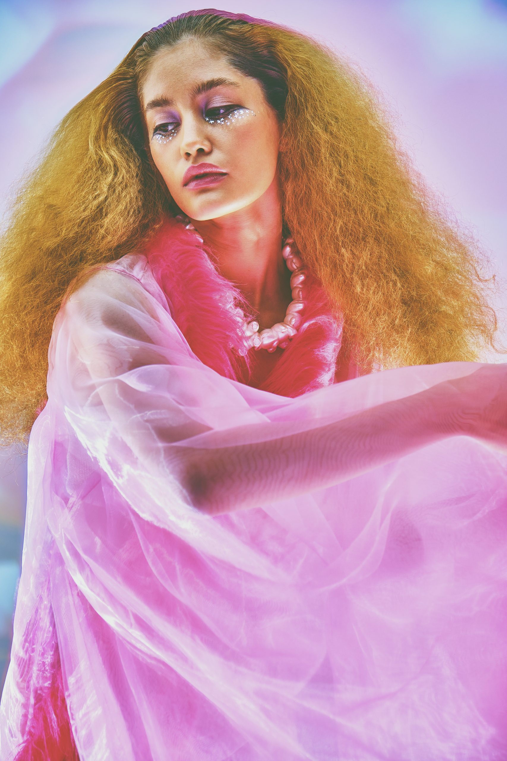 close up shot of White Female Model in sheer pink dress and voluminous hair in front of pink blue background