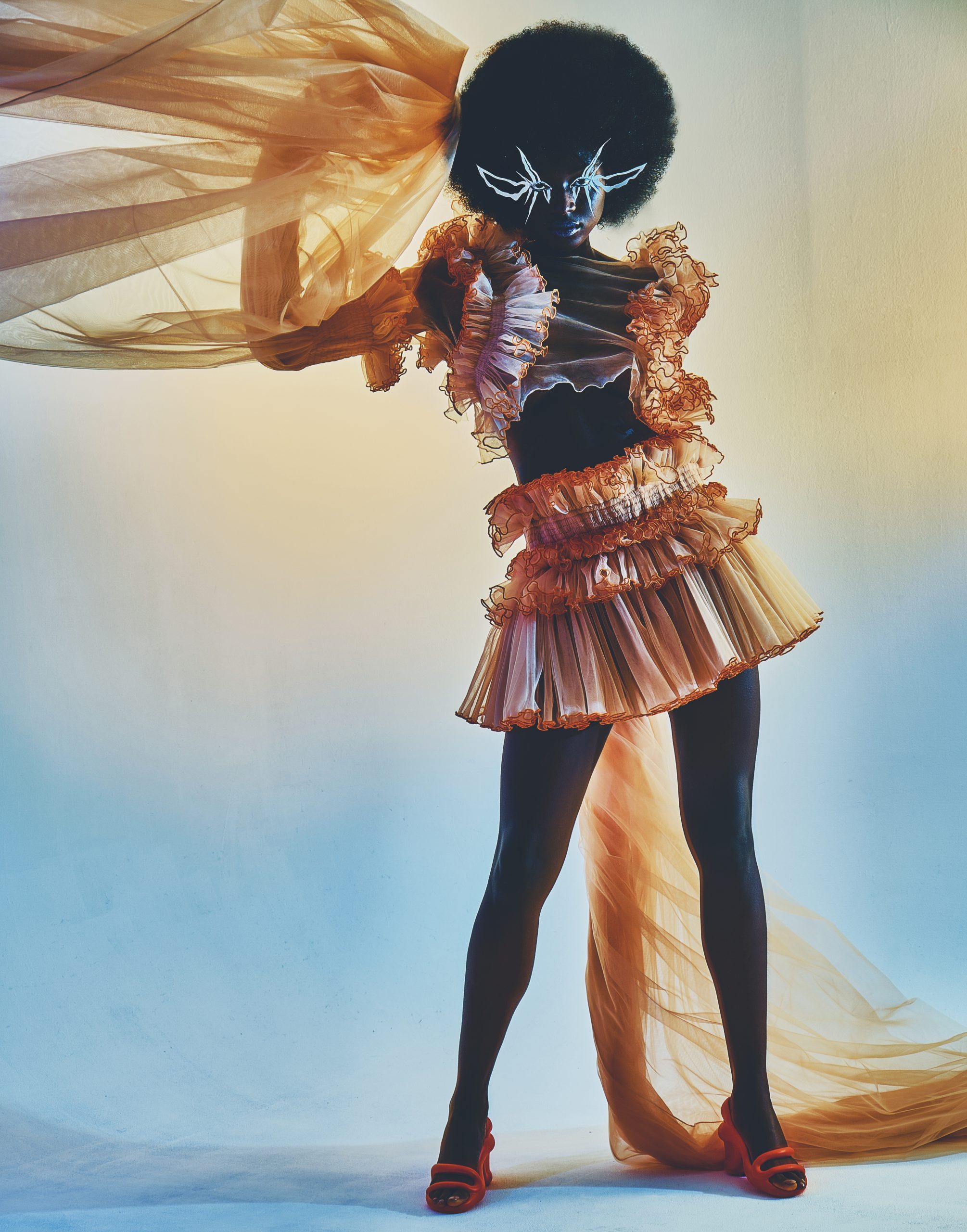 full body shot of dark skin model swinging fabric in upwards motion dressed in ruched orange skirt and top with big afro hair and 3D eyelashes